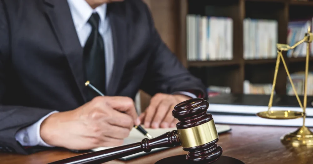 Man in a blue suit writing on paper at a desk with a judge’s gavel prominently in the foreground.